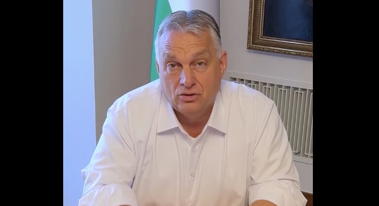 Orbán Viktor kigombolt és feltűrt fehér ingben ül a Karmelita íróasztalánál, háttérben egy magyar záazló