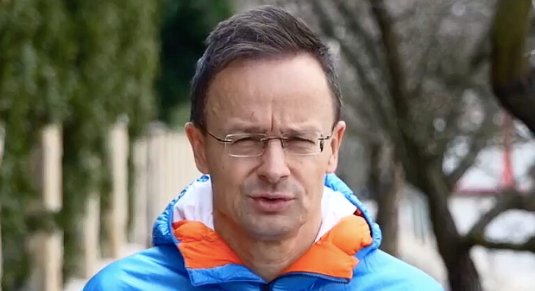 Szijjártó Péter világoskék és narancssárga kabátban a szabadban egy kerítés előtt beszél