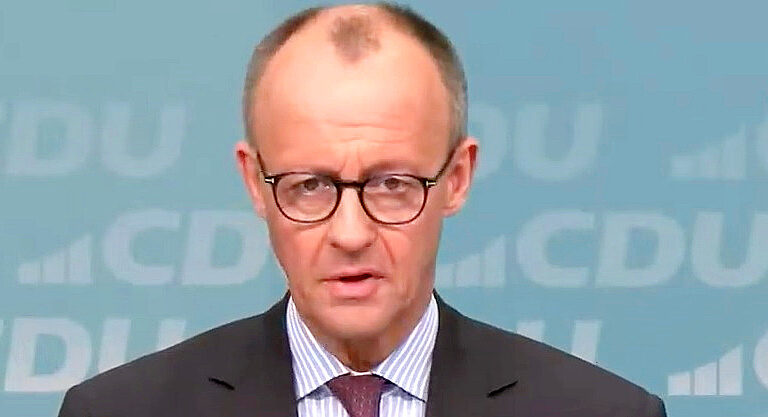 Friedrich Merz fekete keretes, kerek szemüvegben, kék-fehér vertikális csíkos ingben, fekete zakóban, kameréba és gondterhelten néz.