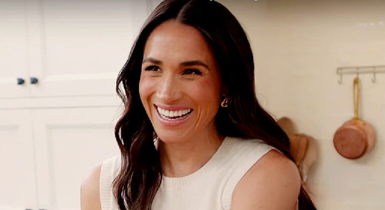 Meghan Markle teli szájjal vigyorog hosszú hajjal, fehér fogakkal, fehér ujjatlan felsőben, miközben enyhén balra néz
