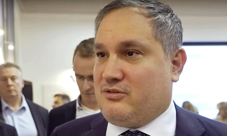 Nagy Márton sötét zakóban, fehlr ingben, sötét nyakkendőben, őszülő hajjal, balra nézve beszél.