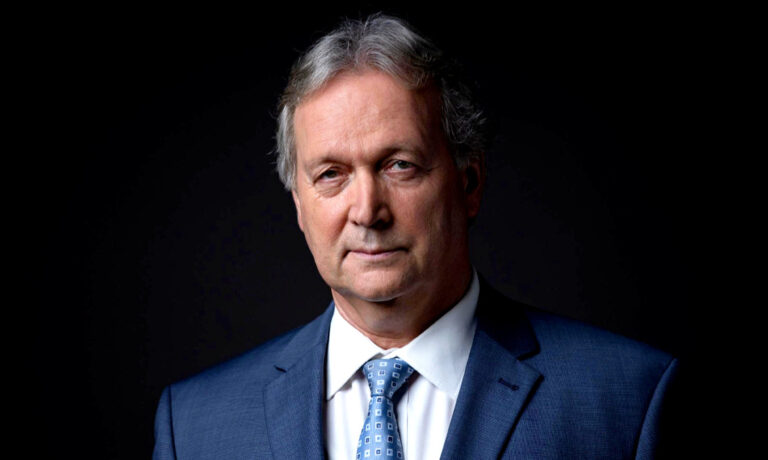 Freund Tamás kék zakóban, fehér ingben és kék nyakkendőben a kamerába néz.