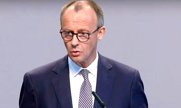 Friedrich Merz sötétkék zakóban, fehér ingben, vörös-fehér nyakkendőben és szemüvegben beszél, miközben kissé gondterhelten enyhén balra néz.