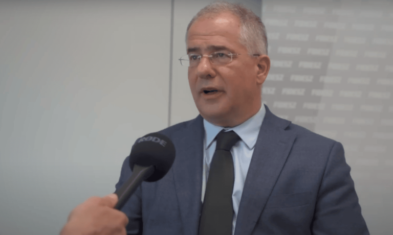 Kósa Lajos szürke háttér előtt szürke zakóban beszél egy mikrofonba