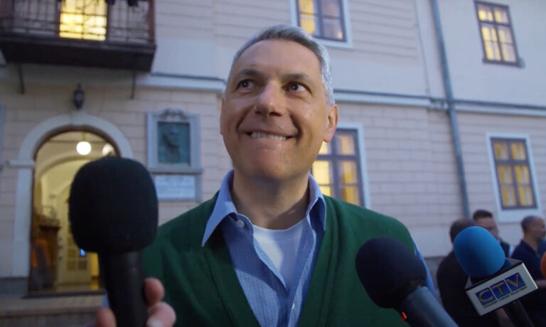 Lázár János harapdálja a szája szélét világoskék ingben és zöld kardigánban miközben mikrofonokkal kérdezik