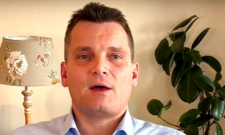 Gaal Gergely világoskék ingben, kameréba nézve beszél egy szobában egy állólámpa, egy növény és egy kanapé elől