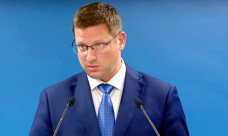Gulyás Gergely kék zakóban, fehér ingben, kék nyakkendőben és szemüvegben enyhén balra nézve, flegmán beszél a mikrofonba.