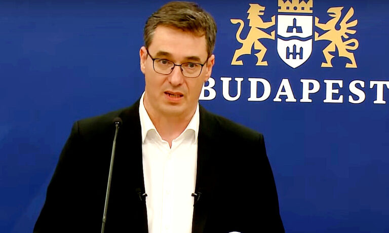 Karácsony Gergely fekete zakóban, fehér ingben, szemüvegben, kócos hajjal mikrofonba beszél, miközben középre néz.
