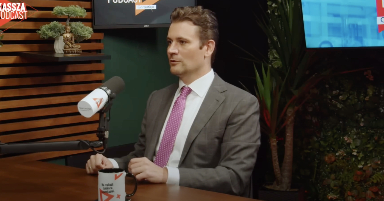 Kisgergely Kornél interjút ad egy mikrofonba beszélve, miközben mögötte egy TV látható