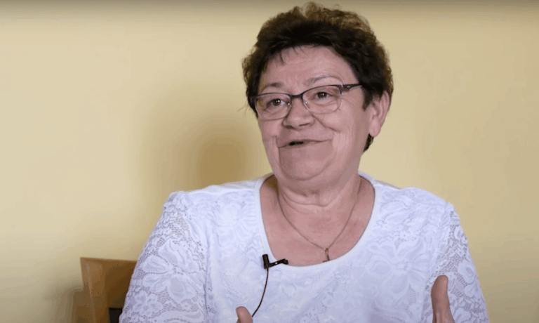 Müller Cecília fehér ruhában mosolyog egy interjú közben drapp fal előtt