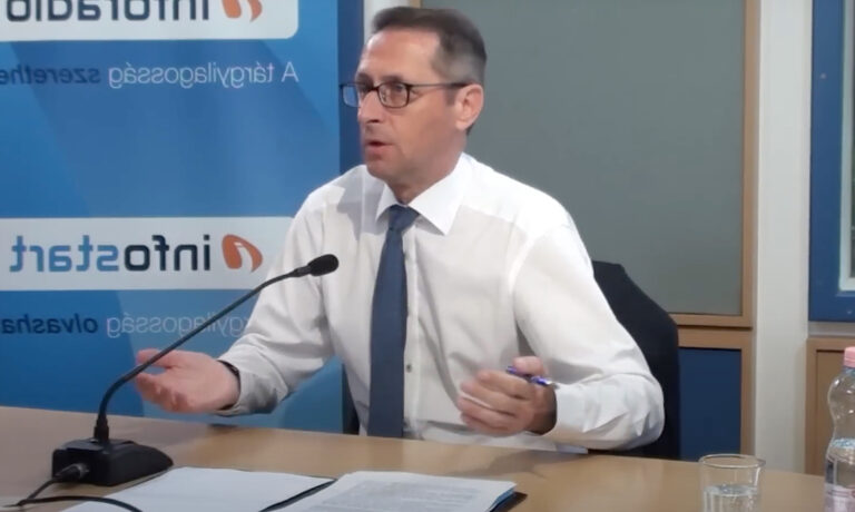 Varga Mihály fehér ingben széttárja a karjait az Inforádió stúdiójában miközben egy kis mikrofonba beszél.