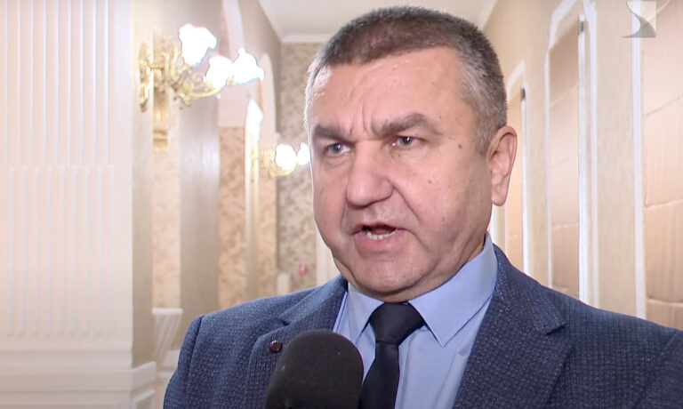 Czomba Sándor interjút ad egy fekete mikrofonba beszélve egy márvány borítású folyosón kék öltönyben és világoskék ingben, fekete nyakkendőben