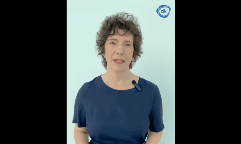 Dobrev Klára kék nyári ruhában egy világos háttér előtt kék DK logóval beszél egy videóban a kamerába