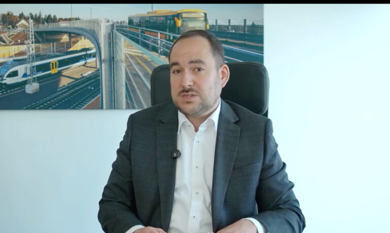 Hegyi Zsolt MÁV-vezér az irodai székében ülve magyaráz a kamerába fehér ingben és sötét zakóban, mögötte a fehér falon egy MÁV vonatot és egy volánbuszt ábrázoló kép