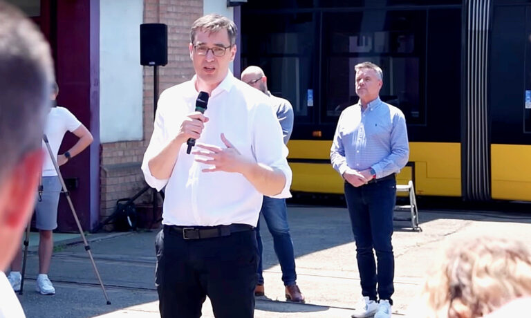 Karácsony Gergely fehér ingben, fekete nadrágban, szemüvegben renkívüli bejelentést tesz a BKV villamoskocsiszínében.