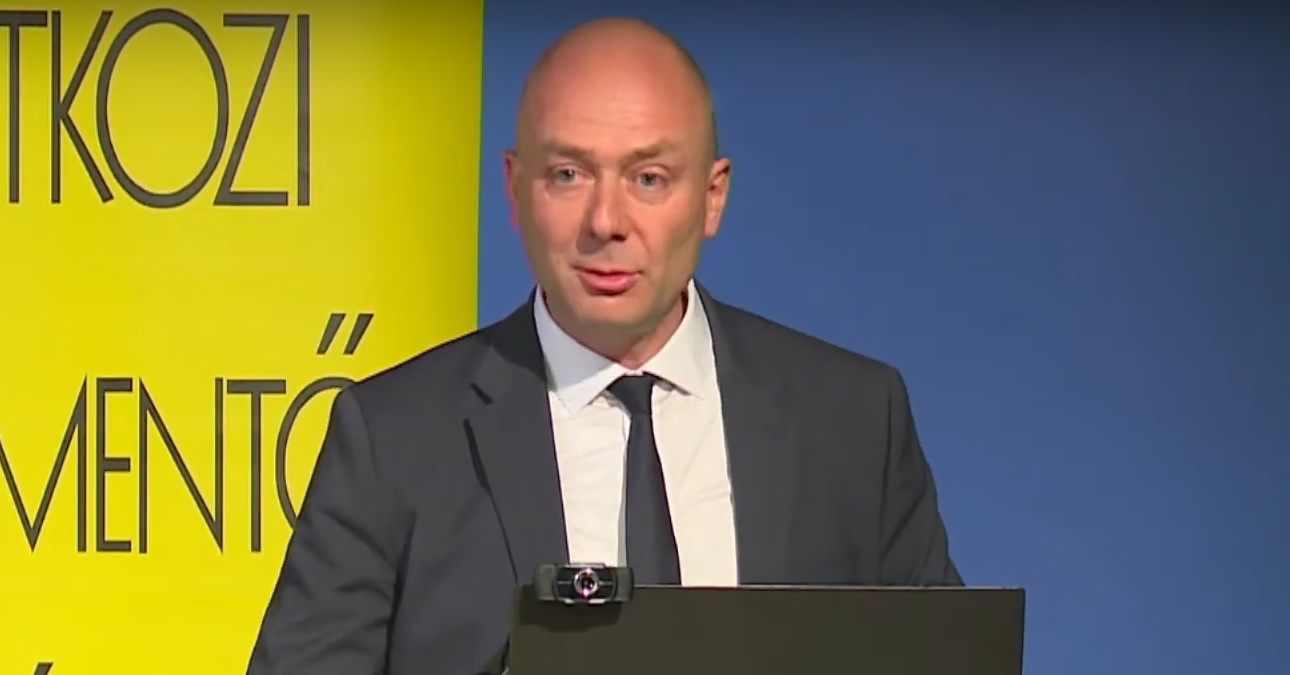 Kozma Ákos szürke zakóban, fehér ingben, fekete nyakkendőben, kopaszon szembenézve beszél.