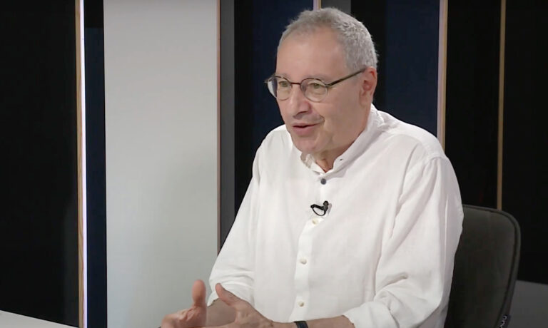 Gábor György fehér ingben beszél egy sötét stúdióban