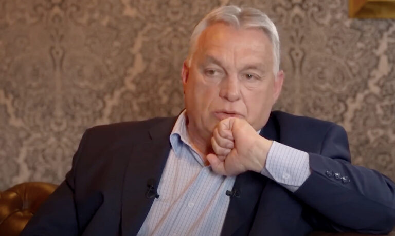 Orbán Viktor egy barna fotelben ülve az öklét a szája elé tartva beszél sötét zakóban és világos kigombolt ingben