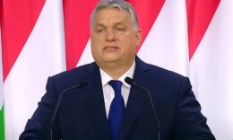 Orbán Viktor zászlók előtt áll sötétkék öltönyben, kék nyakkendőben, mikrofonok előtt