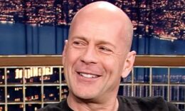 Bruce Willis kopaszon, fekete pólóban, balra nézve mosolyog.