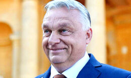 Orbán Viktor sötétkék zakóban, fehér ingben, narancssárga nyakkendőben, balra nézve ijesztően vigyorog.