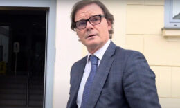 Bánki Erik egy sárga fal előtt szemüvegben sétálva a kamera felé fordul