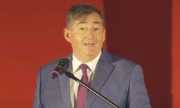 Mészáros Lprinc kék zakóban, fehér ingben, vörös nyakkendőben, vörös háttér előtt mikrofonba beszél.