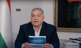 Orbán Viktor egy nemzeti konzultációt szorongat a Karmelitában, a kamerában néz miközben magyaráz a fehér fal és egy kép előtt