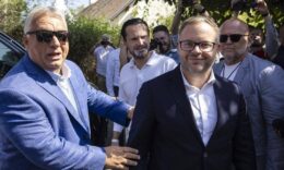 Orbán Viktor és Balázs a napsütésben, előbbi napszemüvegben. Háttérben fotózzák őket a fekete kisbuszból kiszállva, illetve Havasi Bertalan néz.
