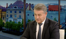 Tasó László egy stúdióban épületes háttér előtt beszél fekete öltönyben és nyakkendőben, fehér ingben