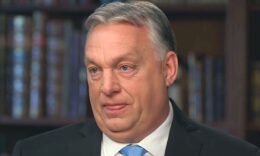 Orbán Viktor megint kinyújtja a nyelvét, könyvespolc előtt ül, fekete öltönyt visel, fehér inget és kék nyakkendőt hord