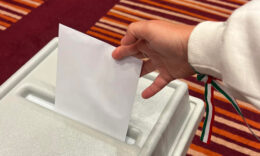 Választás 2024, egy kéz fehér pulcsiban, gyűrűvel az ujján és piros-fehér-zöld karszalaggal a csuklóján bedob egy választási szavazólapot a borítékban egy szavazóurnába. Alatta piros alapon fehér és narancssárga csíkos szőnyeg látható.